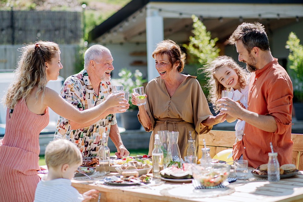 A group of six people, including children and adults, enjoy a meal together outdoors at a wooden table, smiling and raising glasses in a cheerful, sunny setting.