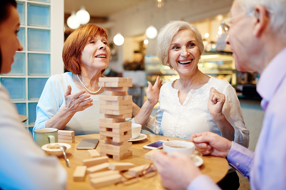 Four older adults sit around a table in a cozy café, smiling and chatting while playing a game of Jenga. Cups of coffee and snacks are on the table, creating a warm and cheerful atmosphere.