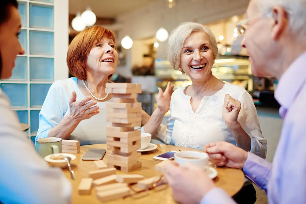 Four older adults sit around a table in a cozy café, smiling and chatting while playing a game of Jenga. Cups of coffee and snacks are on the table, creating a warm and cheerful atmosphere.