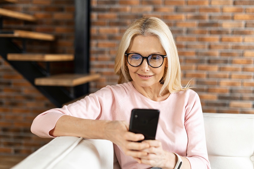 A smiling woman with long blonde hair and glasses sits on a white couch, looking at her smartphone. She is wearing a light pink top and is in a modern room with a brick wall and wooden stairs in the background.