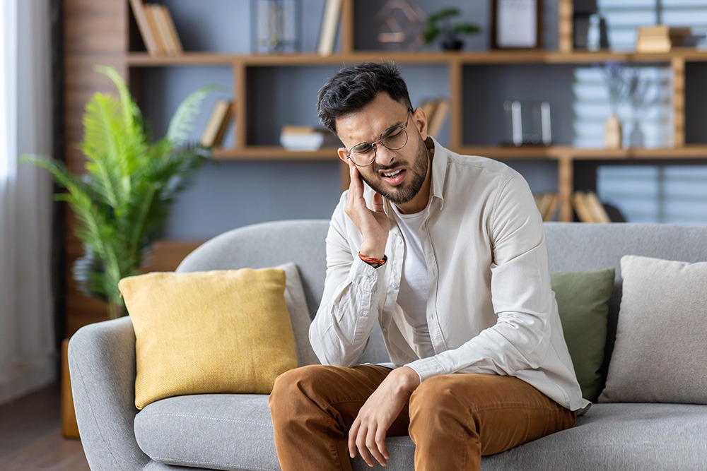 A man sitting on a couch in a living room holds his hand to his ear and appears to be in pain or discomfort. He is wearing glasses, a light shirt, and brown pants. There are bookshelves and plants in the background.