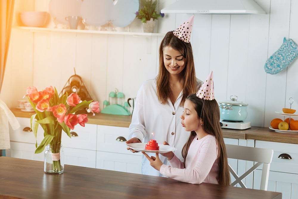 A woman and a young girl wearing party hats celebrate in a kitchen. The woman holds a plate with a small cake and candle while the girl prepares to blow it out. A vase of tulips sits on the table in the foreground.