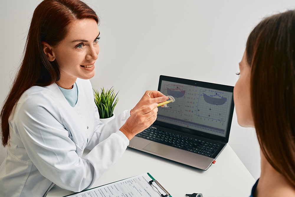 A female audiologist in a white coat explains something to a patient, with a laptop displaying charts and a clipboard on the desk.