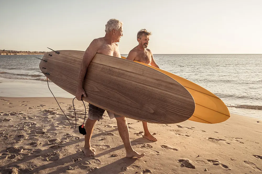 Two older men with gray hair walk barefoot on the sandy beach, each carrying a large wooden surfboard, with the ocean and a clear sky in the background.