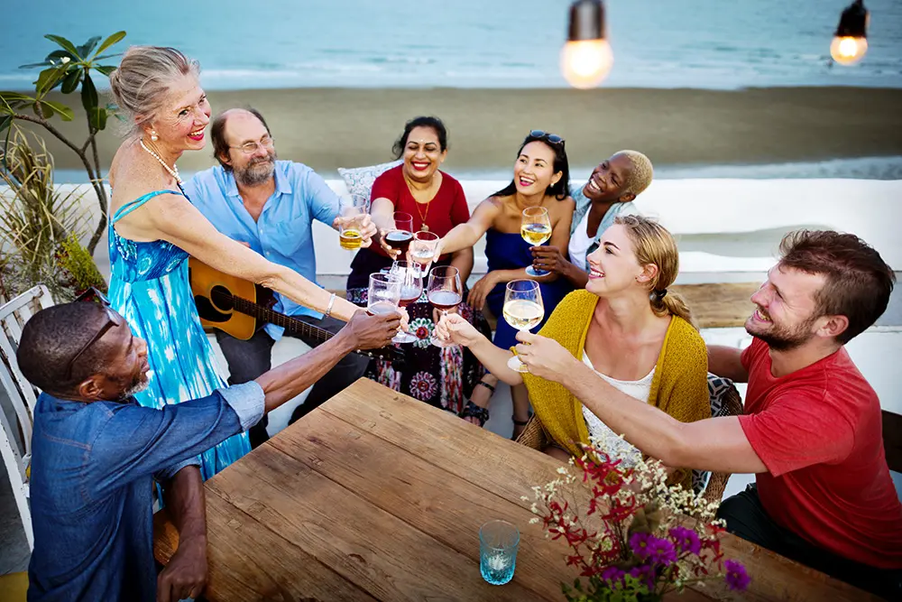 A group of cheerful people sit around a wooden table outdoors by the beach, raising glasses in a toast. One person holds a guitar. The atmosphere is festive and relaxed, with the sea and sandy shore in the background.