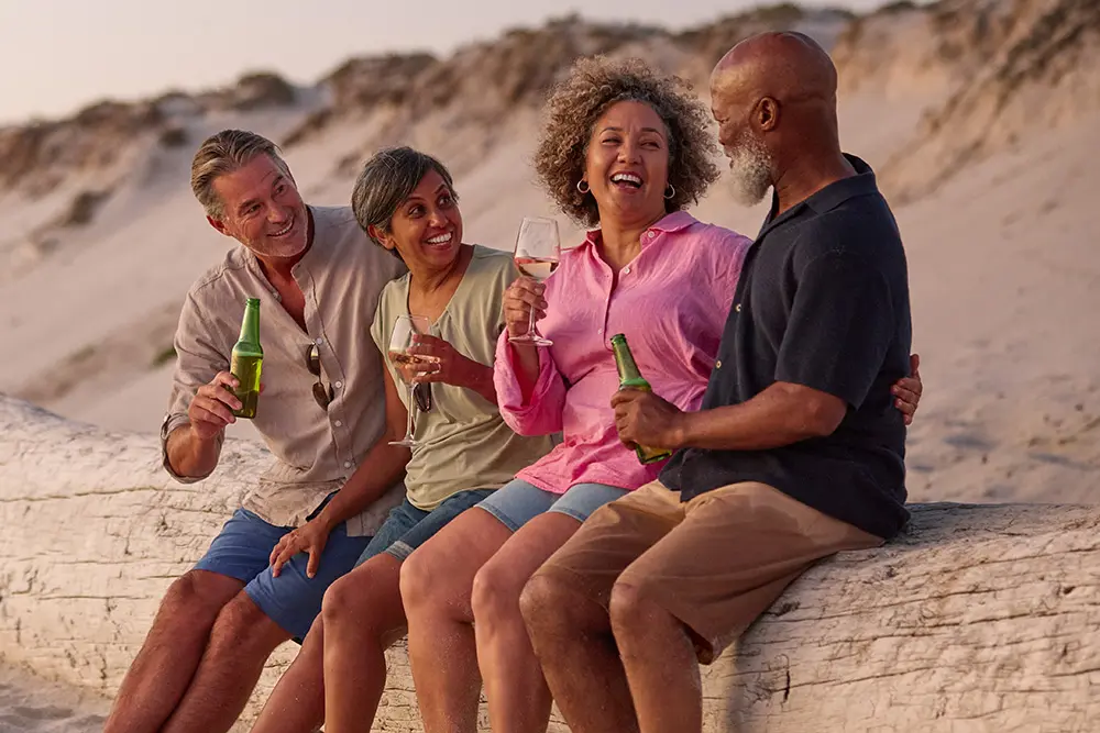 Four adults sit on a large log at the beach, smiling and laughing together while holding drinks. The group appears relaxed and happy, enjoying each others company during sunset. Sandy dunes are visible in the background.